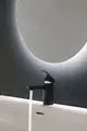 Schwarze Ideal Standard Armatur mit Sensor für Wasserfluss, montiert über einem weißen Waschbecken. Im Hintergrund ist ein beleuchteter runder Spiegel zu sehen.